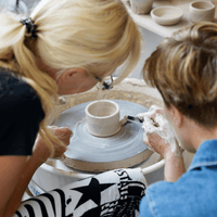 Dwie kobiety siedzące tyłem patrzą na ceramiczną czarkę, którą wytoczyły na kole garncarskim. Jedna trzyma w ręce ceramiczne narządko i obtacza (wykańcza) wotoczone dzieło. W tle widać półki z innymi ceramicznymi pracami.